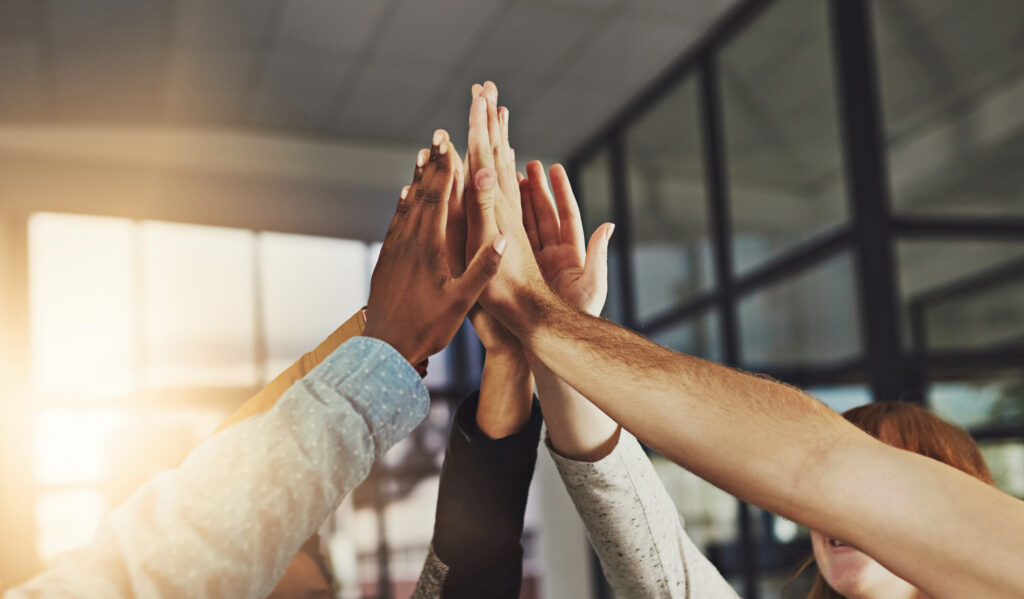 A group of people raising their hands together in a high-five gesture inside a modern office, symbolizing teamwork and collaboration.